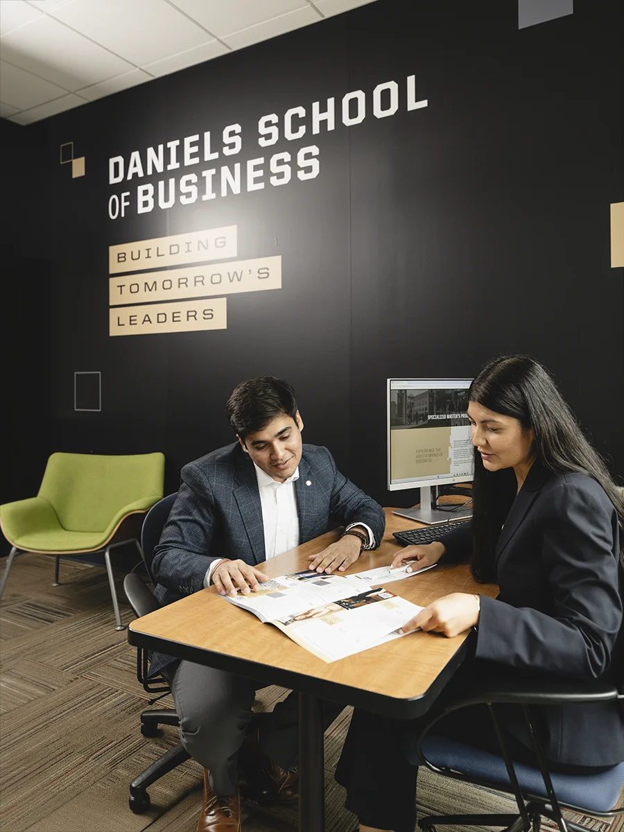 A student and an advisor have a conversation at a desk by a wall that reads Mitch Daniels School of Business