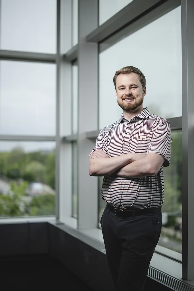 A person with a beard stands with arms crossed near a large window, wearing a striped polo shirt with a logo and dark trousers.