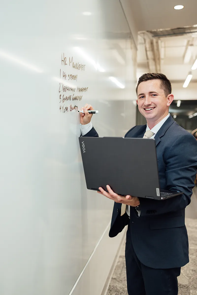 A smiling person in a suit records information from a laptop onto a whiteboard.