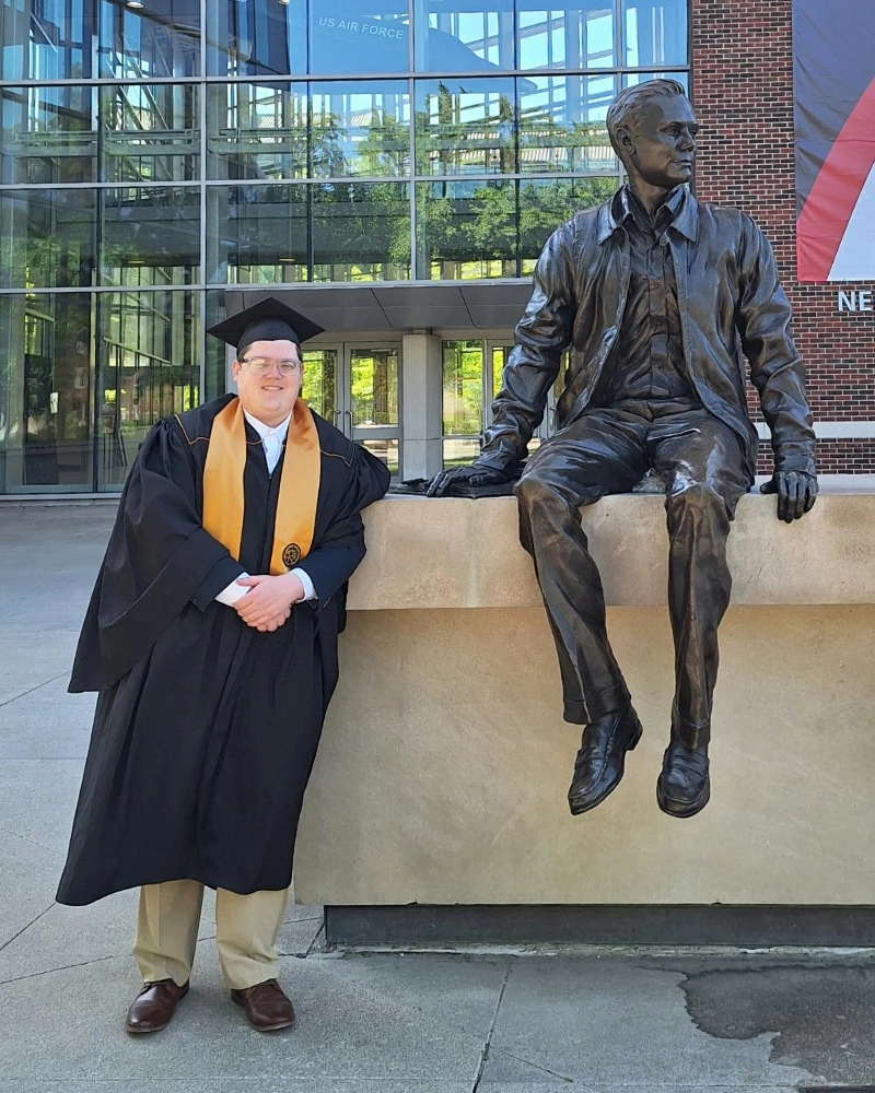 Chris wearing a graduation gown and smiling outdoors next to a statue in Purdue.