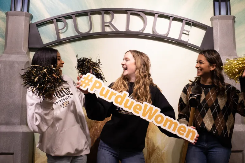 Three laughing students holding pom-poms and a sign that reads #PurdueWomen